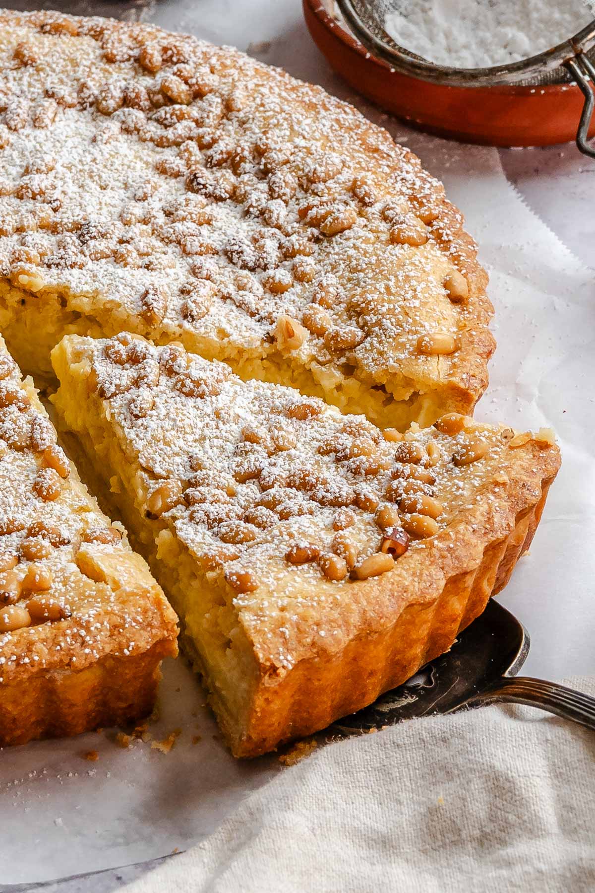 Close-up of a sliced Italian custard tart, showing the creamy yellow filling, golden crust, and pine nuts dusted with powdered sugar, with a cake server underneath the slice.