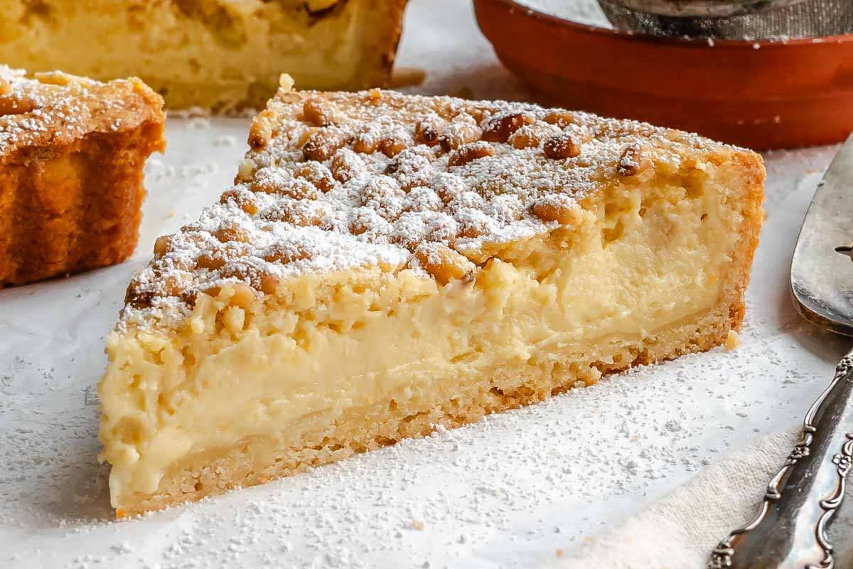 Side view of a single slice of torta della nonna, showing the layers of buttery shortcrust and smooth vanilla custard under a pine nut and powdered sugar topping.